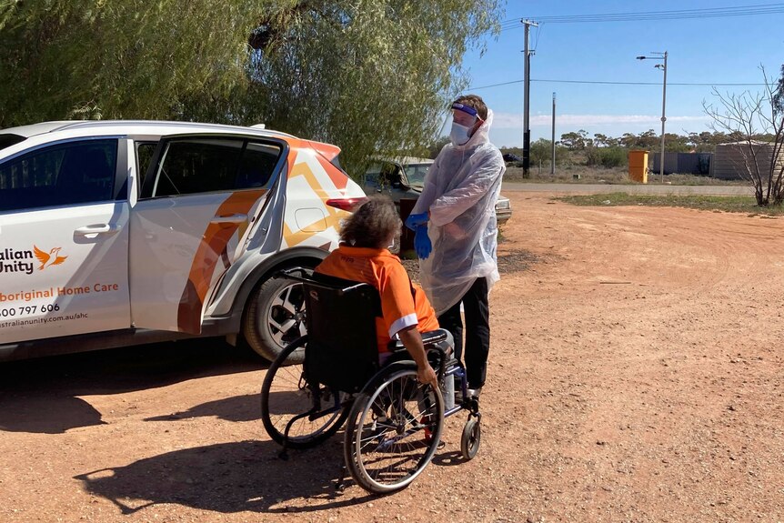 A man in Pesonal Protective Equipment helps an elderly indigenous woman in a wheelchair into a car