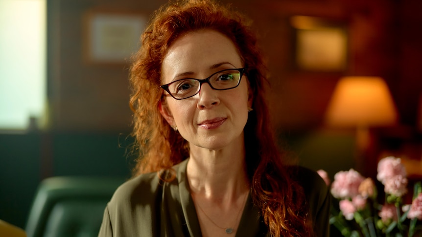 White woman with fiery red curly hair wears dark glasses and an olive green blouse in a warmly-lit room.