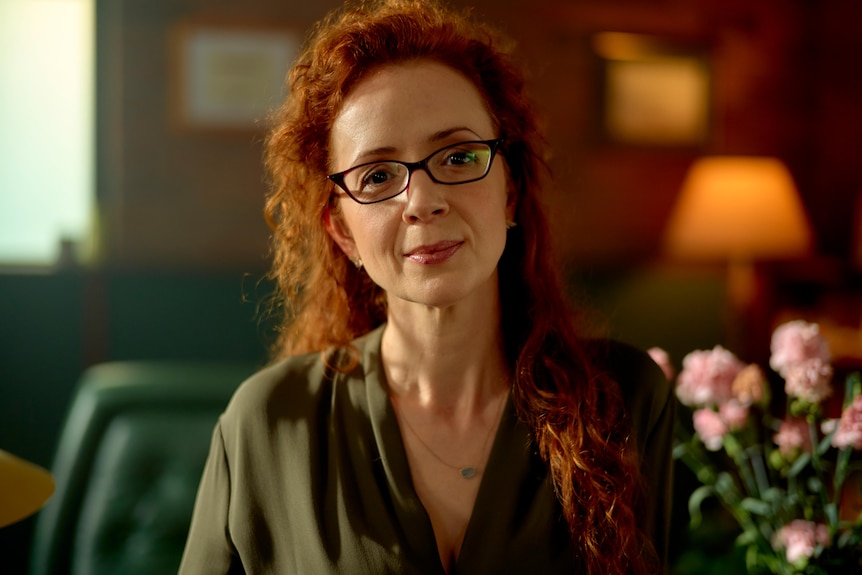 White woman with fiery red curly hair wears dark glasses and an olive green blouse in a warmly-lit room.