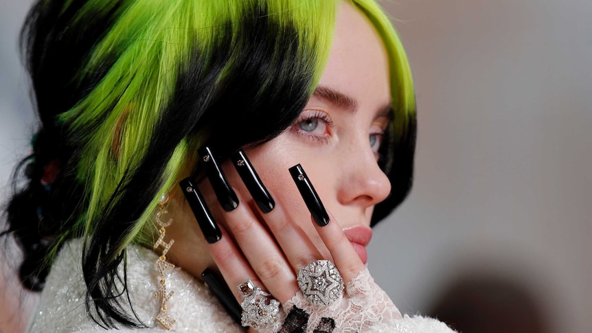 A close-up shows Billie Eilish holding up her long black nails next to her face.