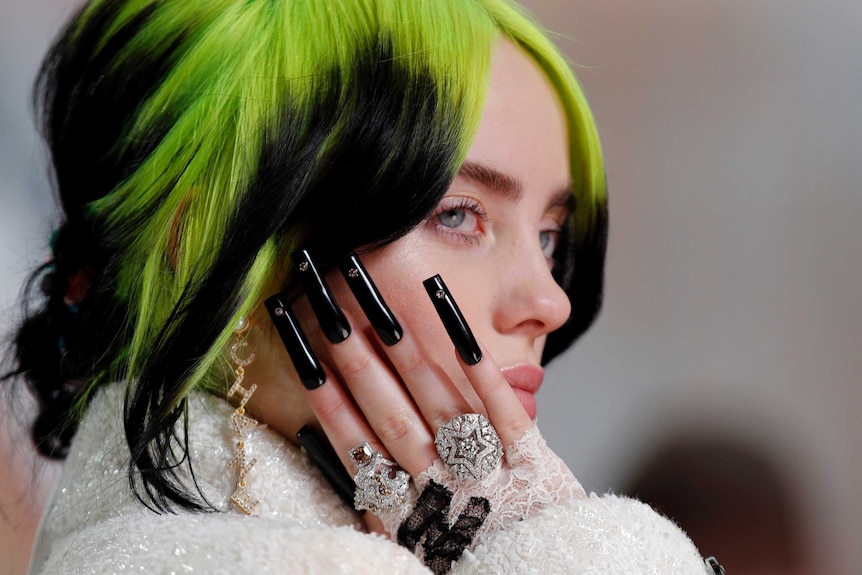 A close-up shows Billie Eilish holding up her long black nails next to her face.