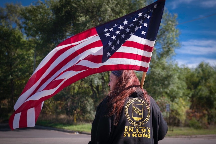 A woman holding a US flag stands with her back to the camera, her jacket with 'Union Strong' written across it 
