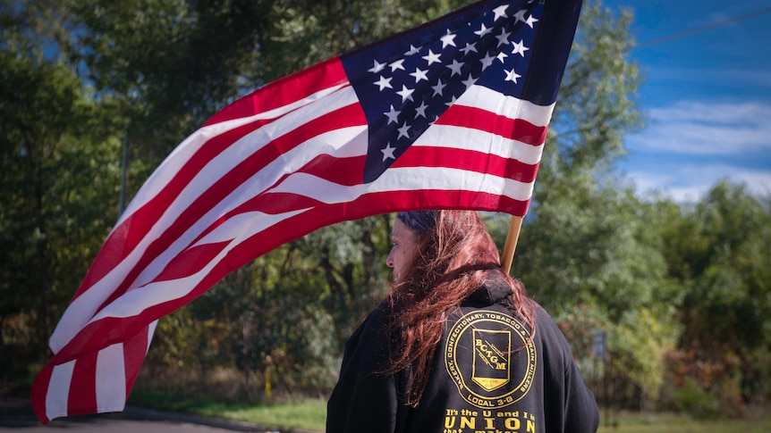 A woman holding a US flag stands with her back to the camera, her jacket with 'Union Strong' written across it 