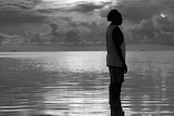 A black and white image of Daniel Billy in silhouette, standing in the ocean shallows.
