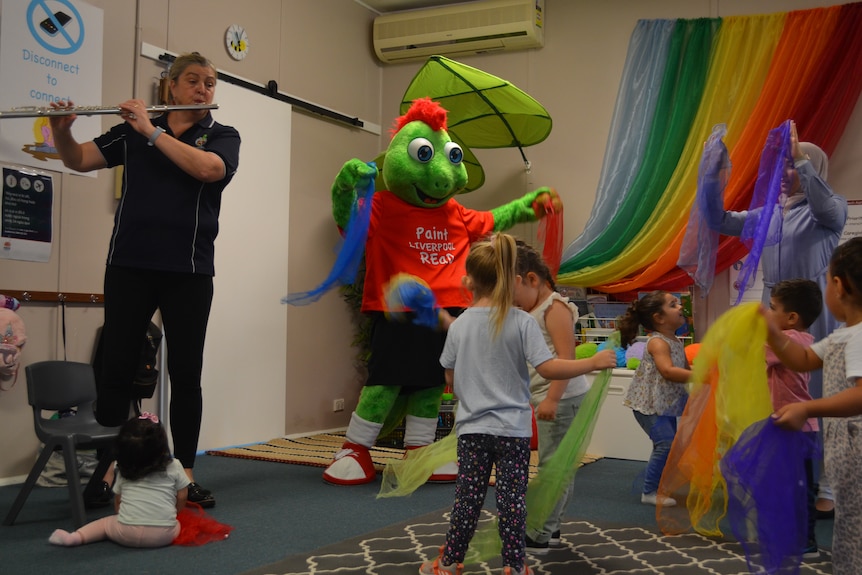 A gecko mascot dances with children holding rainbow streamers while a woman plays the flute