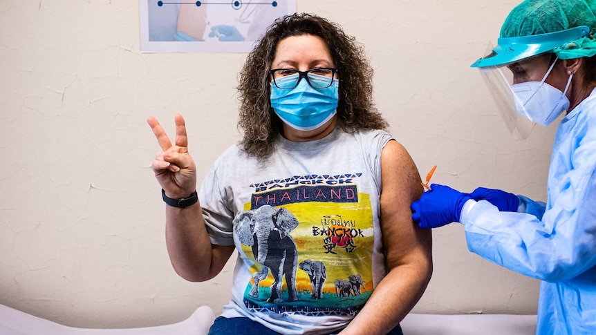 A woman getting a vaccine in her left arm makes a V sign with her right hand