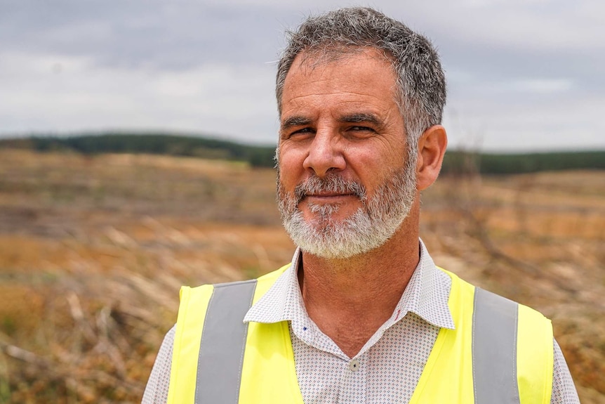 A man with a grey beard wearing a high vis vest stands on a forestry plantation burnt down by a bushfire.