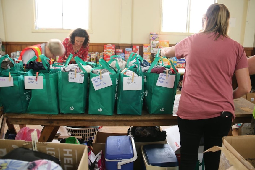 Green shopping bags filled with children's clothes line a table. Groceries sit on another table. Three women sort items.