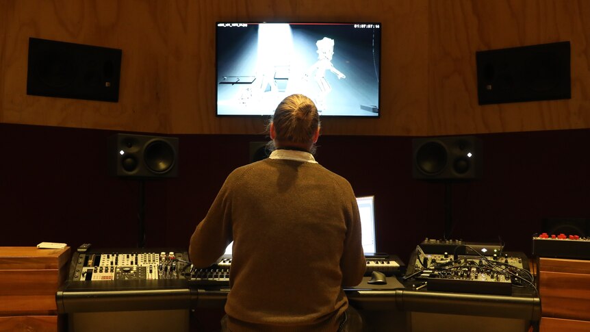 view of man from behind sitting at studio mixing desk with screen above him with animation on it