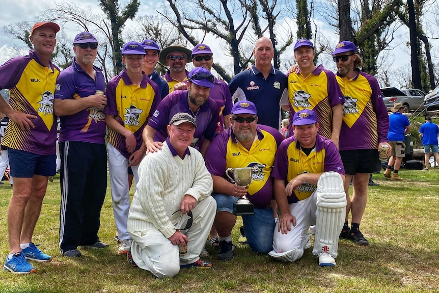 A men's cricket team huddles around a trophy after winning a match