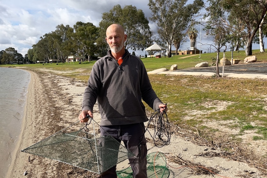 A man holding a pyramid shaped fishing net. There is a lake and sandy grass behind him.