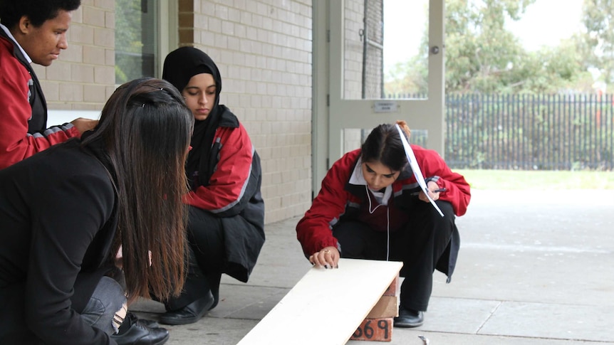 Two students and Lori Lu watch Keharn Hudson release a marble at the top of a ramp outside a classroom.