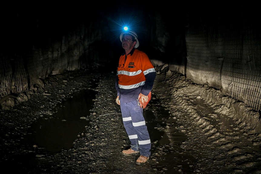 A man wearing orange overalls and a helmet underground with a light on it.