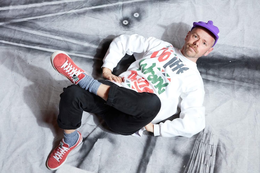 40-something man wearing purple cap, white sweater, black pants, red trainers, lying on ground in pose, photographed from above.