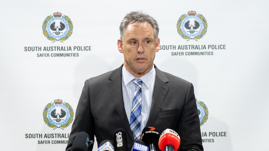 A man wearing a suit stands behind media microphones and in front of a banner with SA Police logo