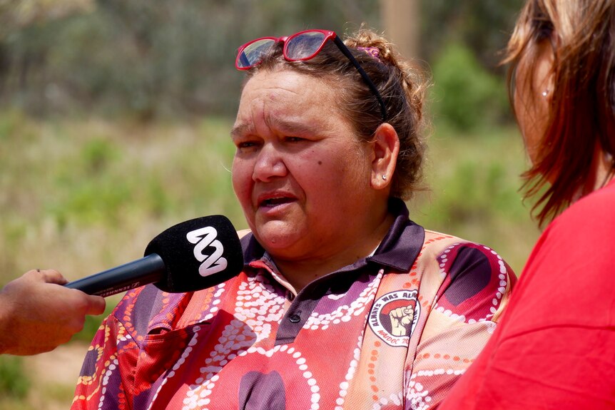 An indigenous woman wearing a red shirt and glasses speaking into an ABC microphone. 