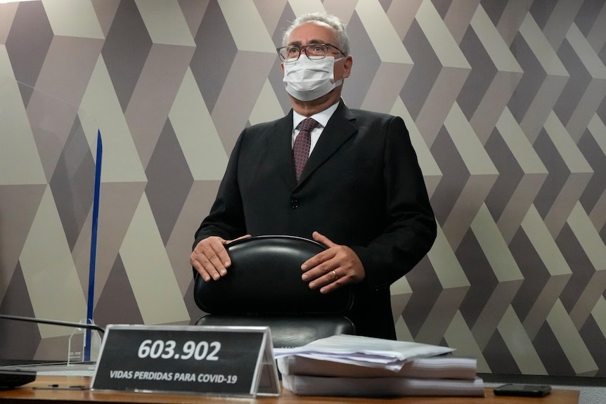 Man stands behind a chair wearing a suit and a white face mask.