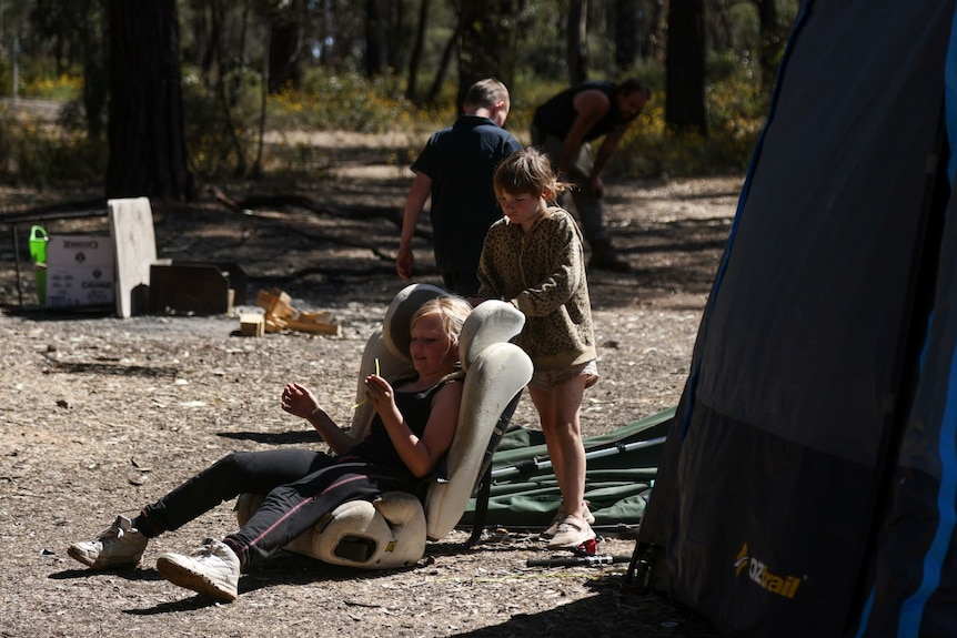 A child sits on a car seat in a campsite, with another child behind the seat.