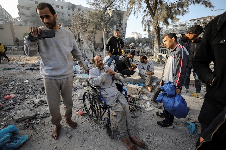 A man sitting in a wheelchair surround by others in an area filled with rubble