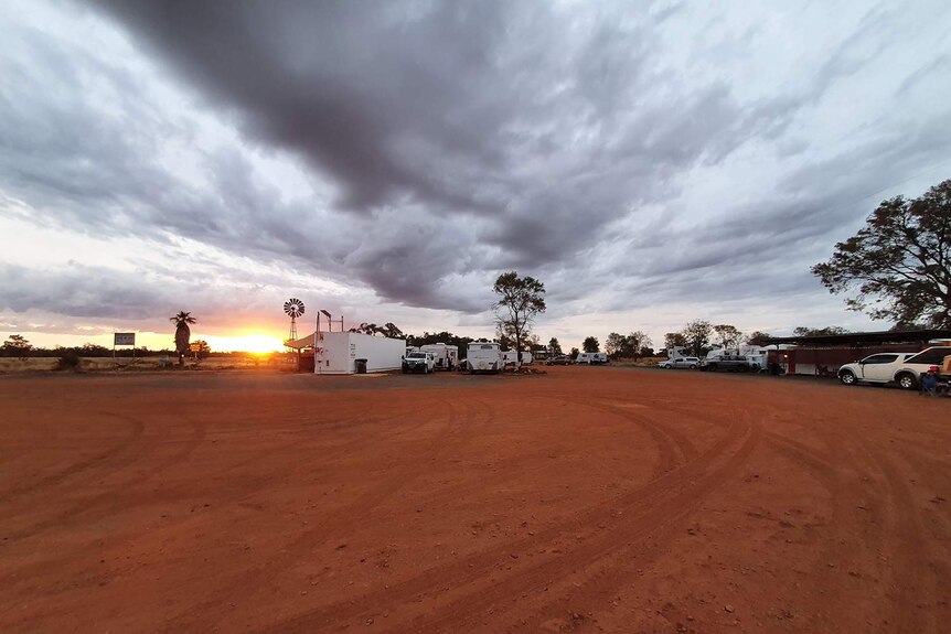 The setting sun shines between an overcast sky and red dirt caravan park