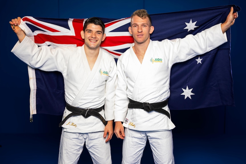 Two men in judo outfits pose holding an Australian flag behind them