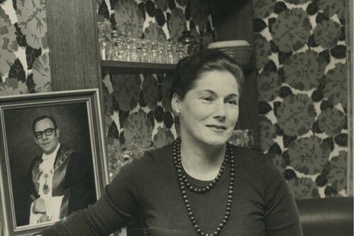 Black and white photo of Pam Stone in a lounge room circa 1977 with a portrait of Les Stone in the background