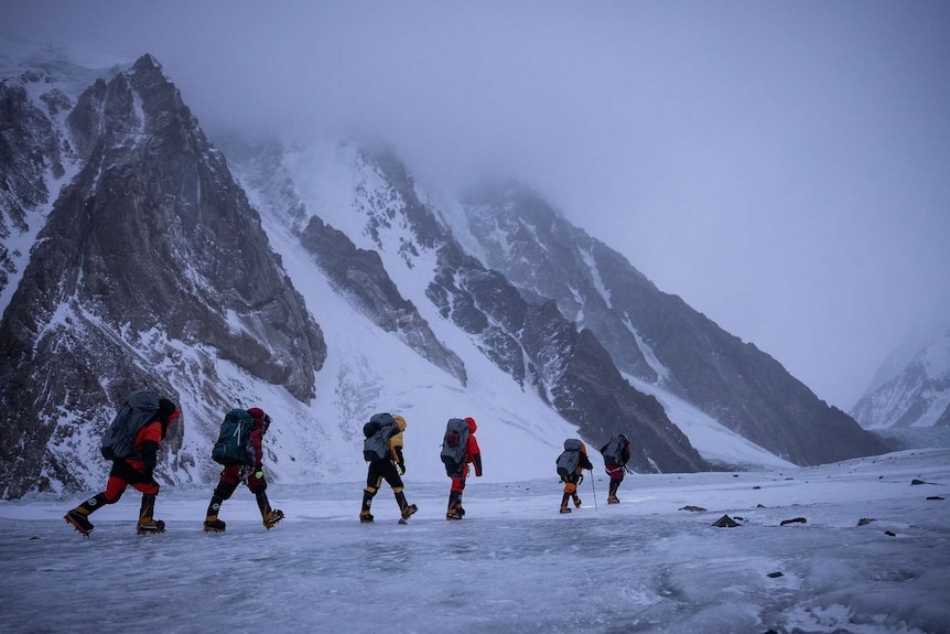 A team of climbers in bright jackets walk in front of the face of K2
