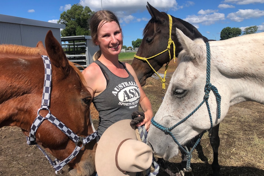 A woman, surrounded by horses,wears a grey singlet, holds a hat and smiles.