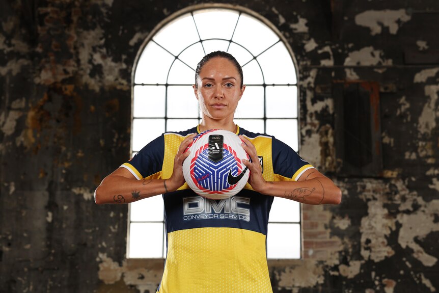 A woman wearing yellow and blue holds a soccer ball to her chest with a window in the background