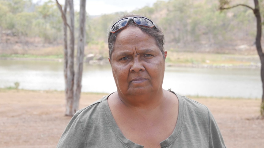 A woman looks at the camera. She has short dark hair and is wearing a green top. The background features a dam and trees.