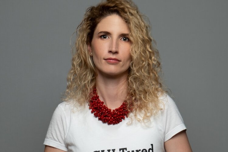 A woman with blonde curly hair and a red necklace, looking seriously at the camera.