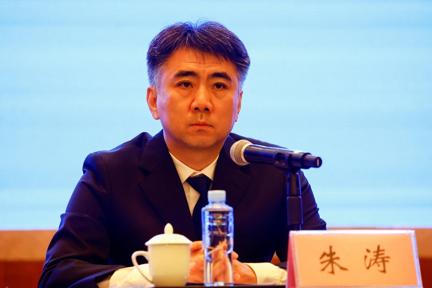 Man in a suit sitting in front of a microphone speaking to a news conference.