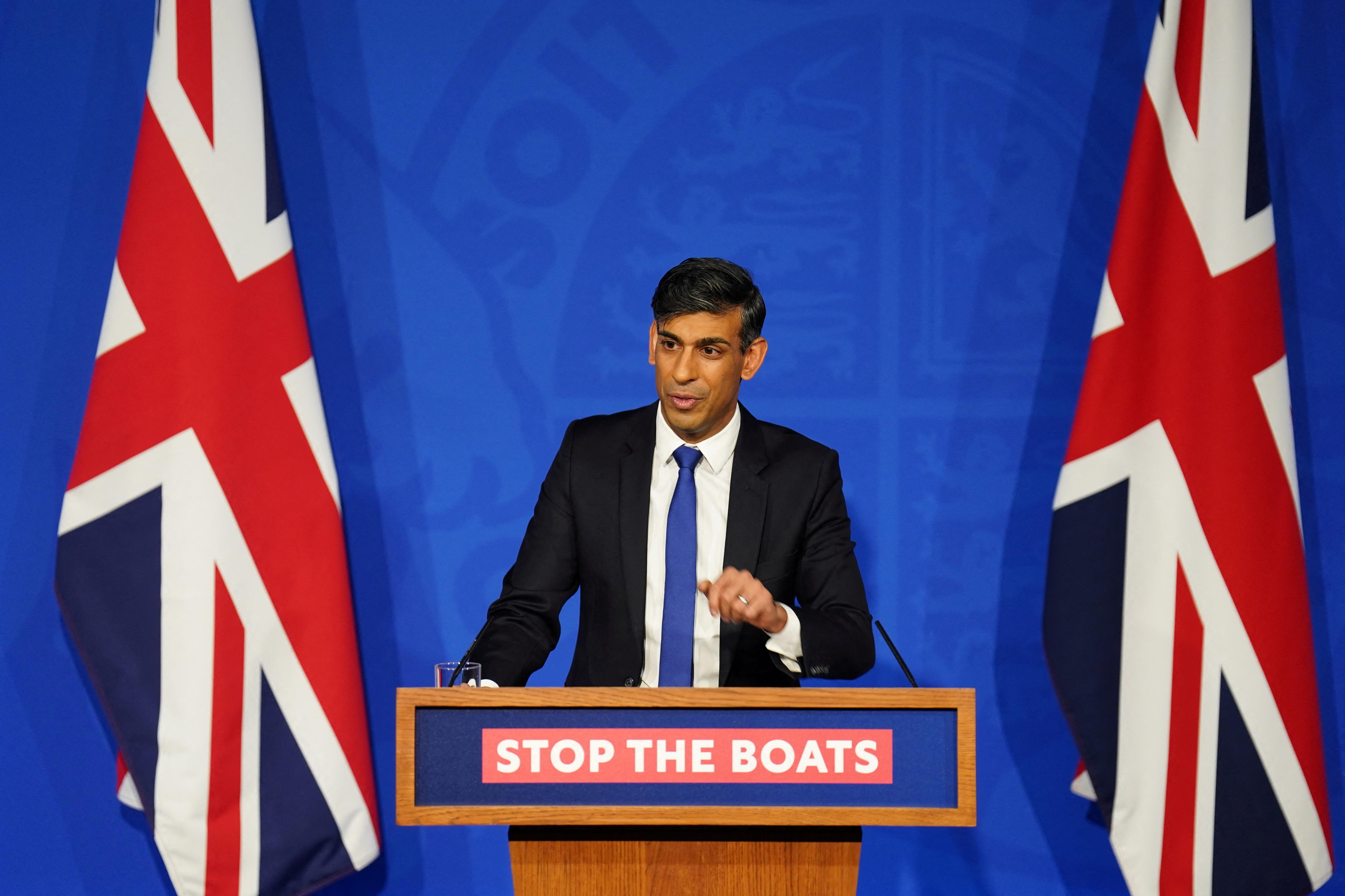 A man in a suit stands behind a podium emblazoned with 'STOP THE BOATS', between two UK flags.