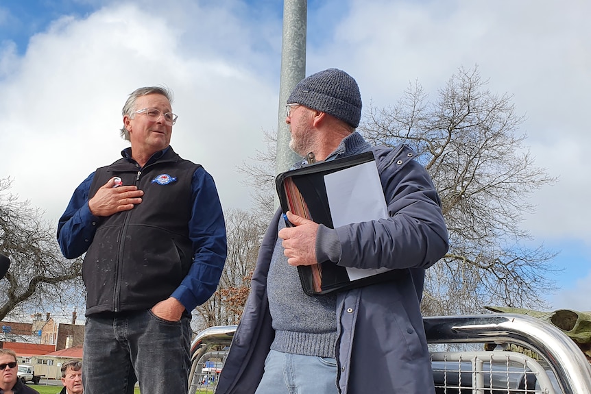 Two men stand on the back of a ute, speaking.