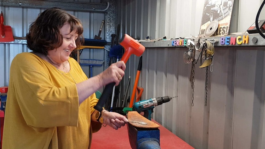 A woman with short brown hair and a yellow knitted sweater flattens a spoon on an anvil by using a hammer.