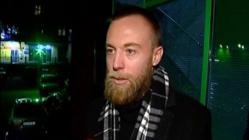 A bearded man wearing a dark coast and a scarf talks to reporters outside a police station.