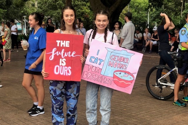 Two teenagers hold signs saying"The future is our" and "I eat gender norms for breakfast".