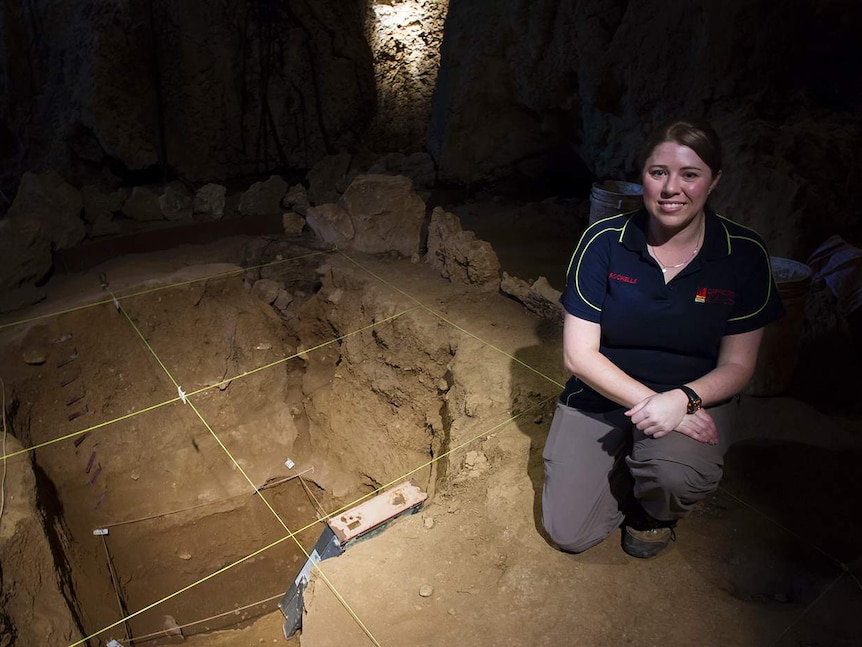 A woman kneels on one knee in a cave, smiling, in front of a dug out pit with a grid of string above