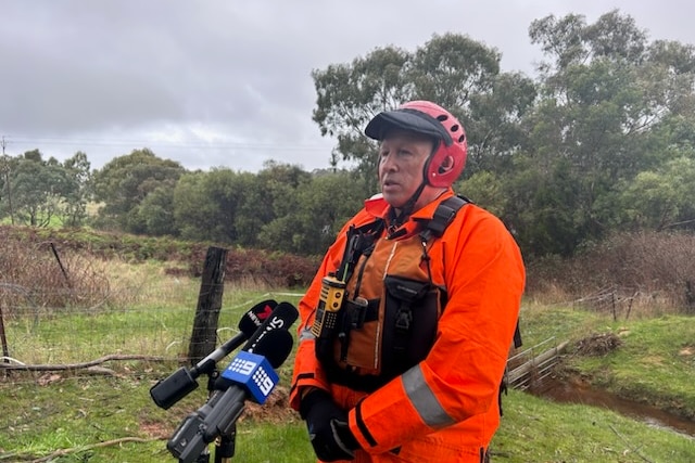 A man wearing an orange jumpsuit and helmet stands in front of media microphones