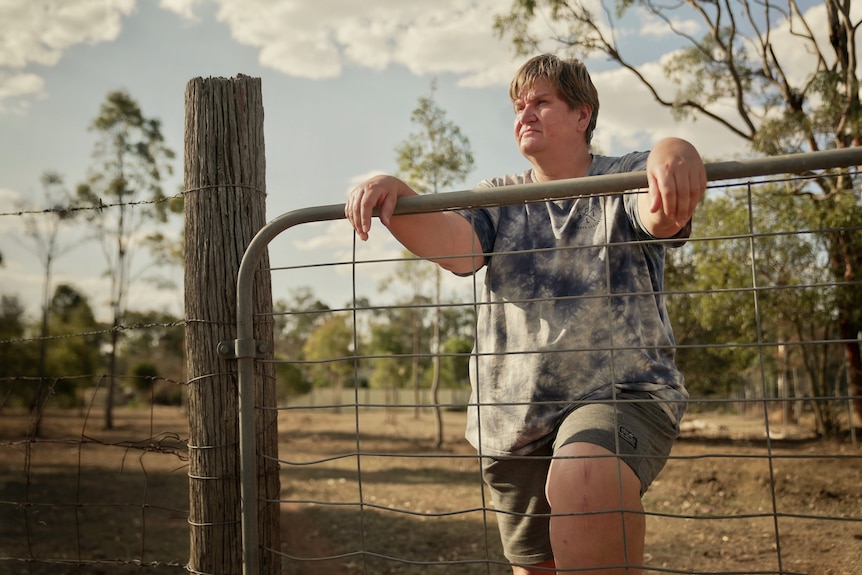 A woman wearing shorts and a T-shirt standing in front of a fence with a pensive expression.