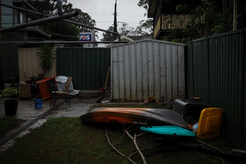 A kayak, branches and other flood debris on grass in front of a backyard shed