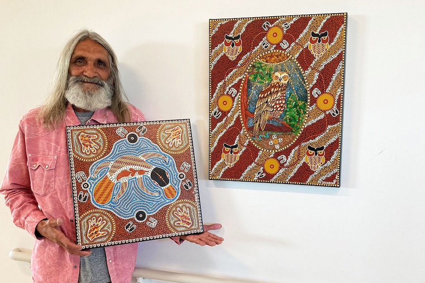 Indigenous artist Stanley Geebung smiles as he stands with two of his artworks.