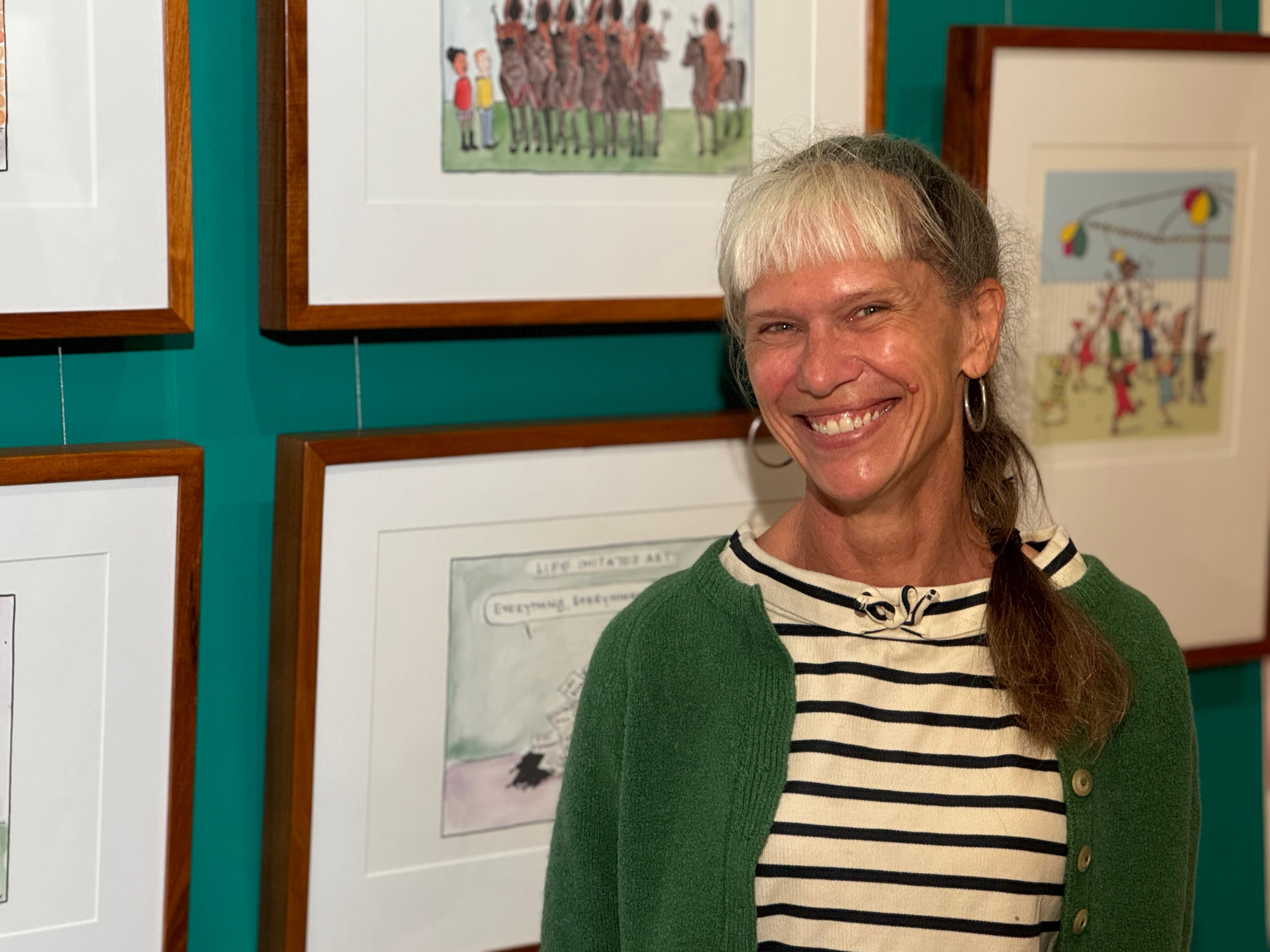 A woman in a green cardigan and stripy top stands in front of some framed cartoons.