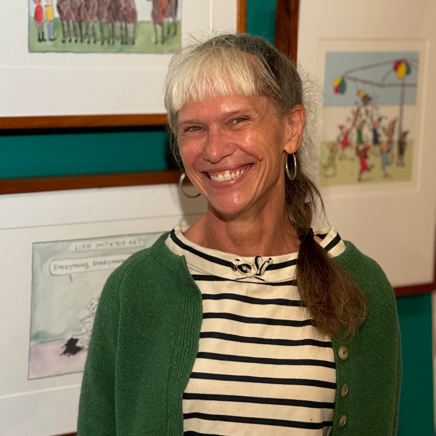 A woman in a green cardigan and stripy top stands in front of some framed cartoons.