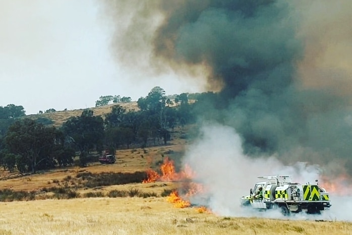 A firetruck shrouded in smoke next to a grassfire with unburnt trees in the background, a helicopter is also in the air.