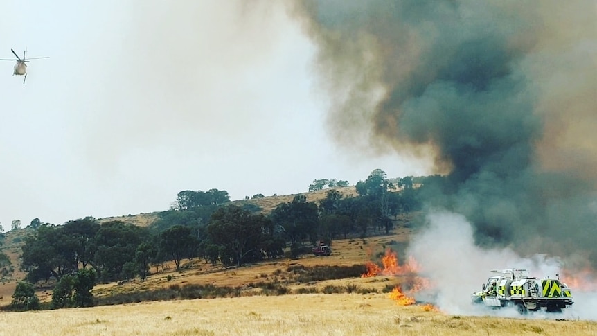 A firetruck shrouded in smoke next to a grassfire with unburnt trees in the background, a helicopter is also in the air.