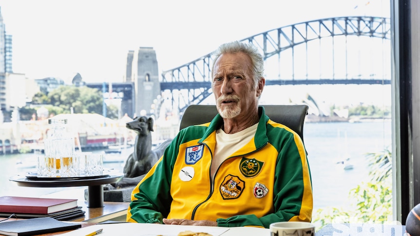 Bryan Brown dressed in a green and gold tracksuit sitting in an office, the Sydney Harbour Bridge behind him
