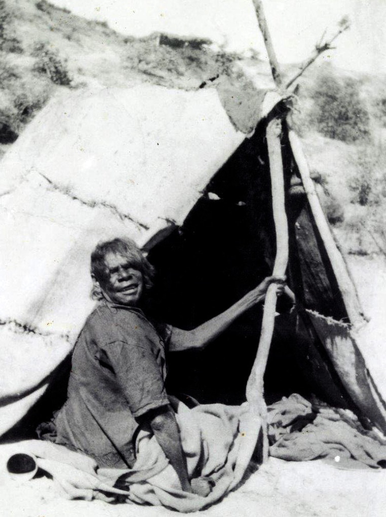 An older woman sits in front of a canvas tent supported by sticks.