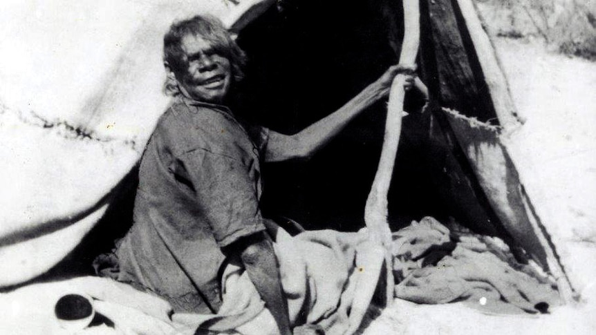 An older woman sits in front of a canvas tent supported by sticks.