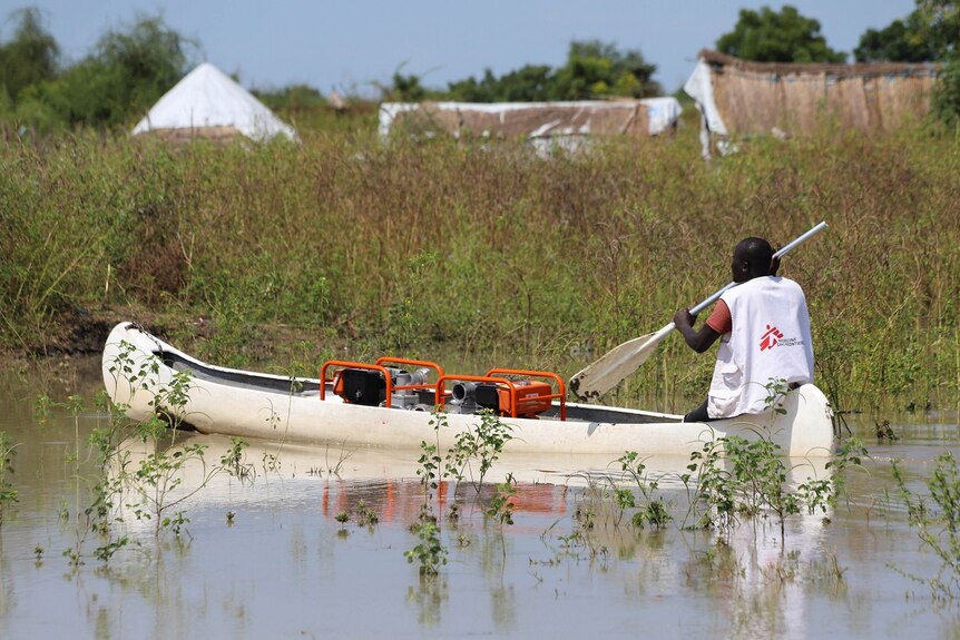 A man wearing a vest with the Medecins Sans Frontieres logo on the back paddles a canoe through floodwaters.